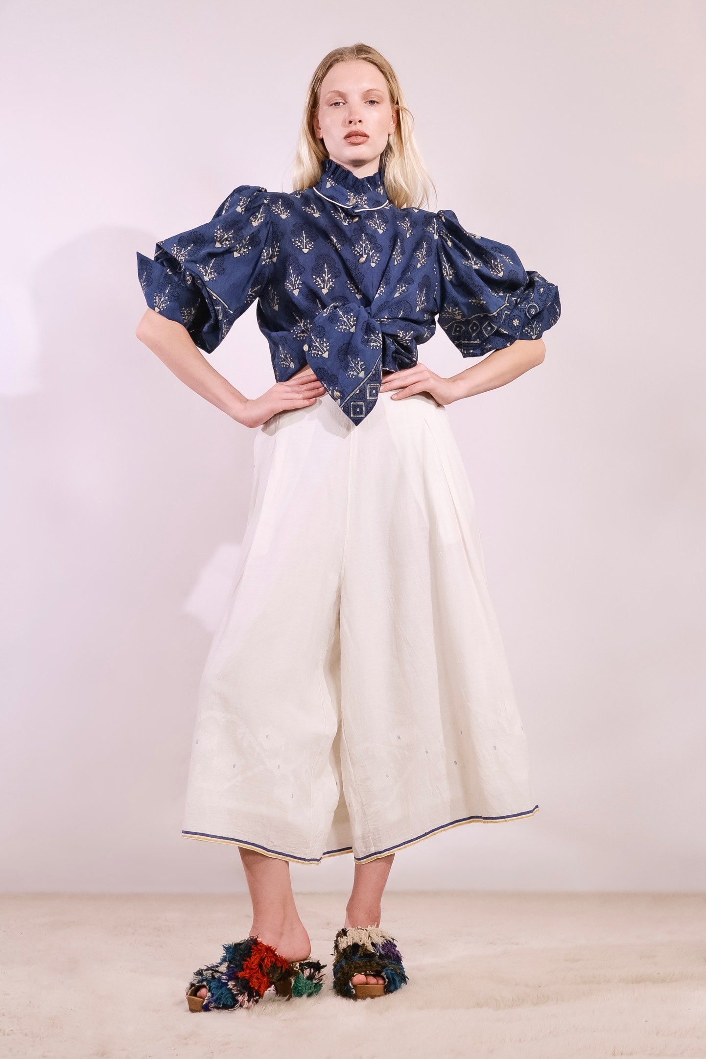 Model is wearing the Amber 59 handloomed pyjama trousers by Injiri — wide-leg organic cotton pants in crisp white with cobalt and yellow jamdani weaving along the hem, featuring an elasticated back waistband and side pockets.