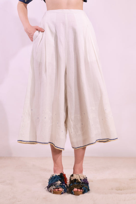 Model is wearing the Amber 59 handloomed pyjama trousers by Injiri — wide-leg organic cotton pants in crisp white with cobalt and yellow jamdani weaving along the hem, featuring an elasticated back waistband and side pockets.