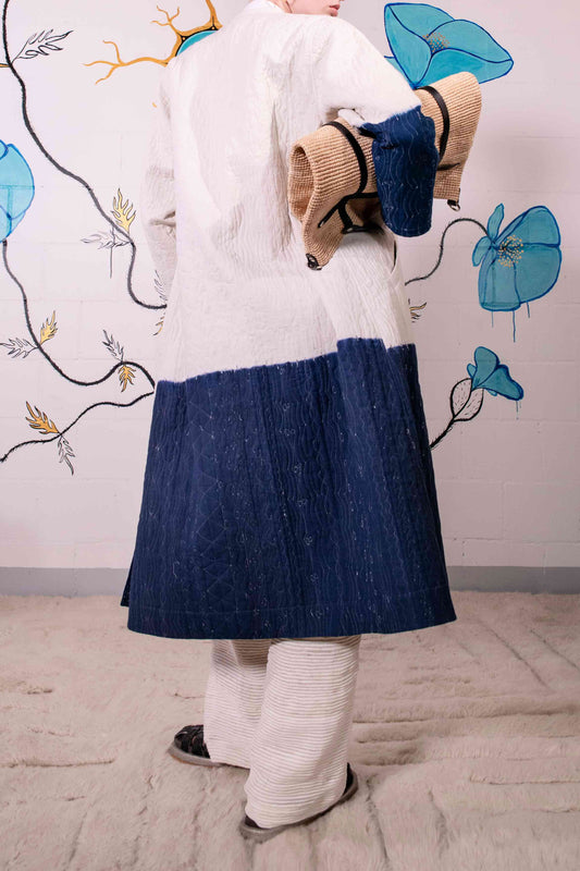 Model is wearing a reversible quilted cotton coat by Injiri, featuring a tie-dye design in off-white and deep indigo, with a split rounded collar, contrast buttons, and blue madras check lining.