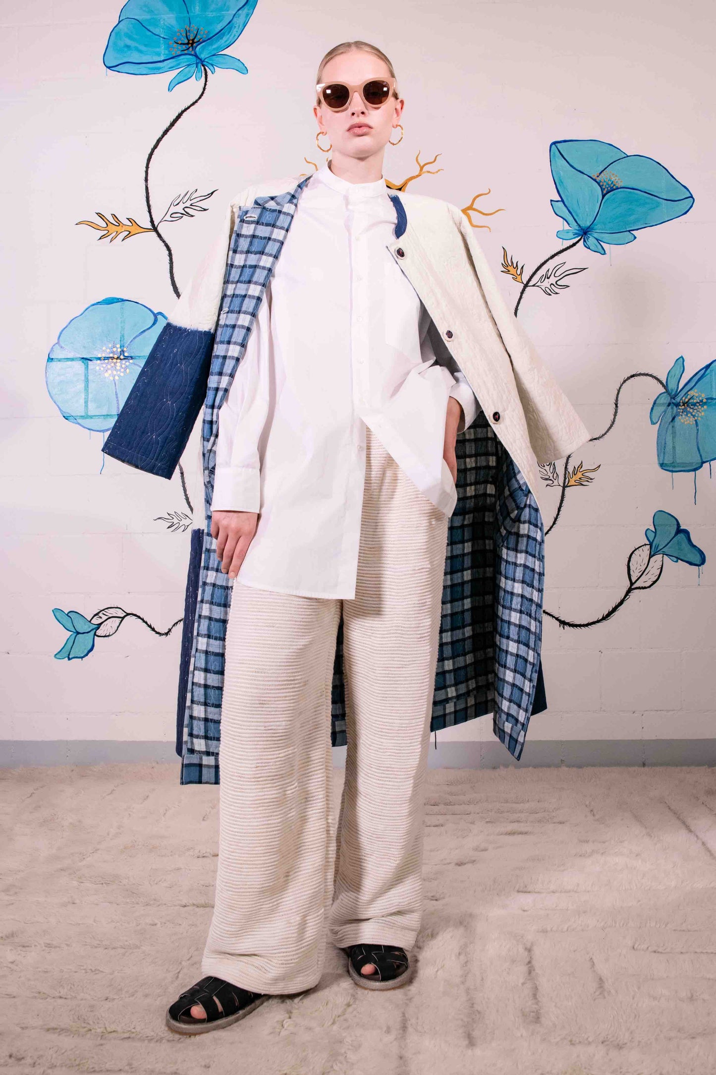 Model is wearing a reversible quilted cotton coat by Injiri, featuring a tie-dye design in off-white and deep indigo, with a split rounded collar, contrast buttons, and blue madras check lining.