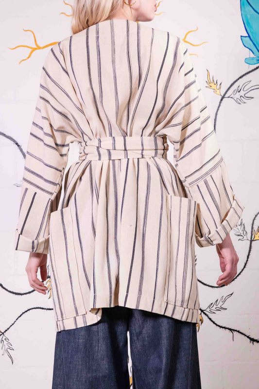 Model is wearing a midi-length, handwoven and hand-printed Benin cotton kimono by Atelier V.O, featuring traditional Dogon and Nagô-inspired patterns, a straight cut, drawstring closure, and wooden buttons.