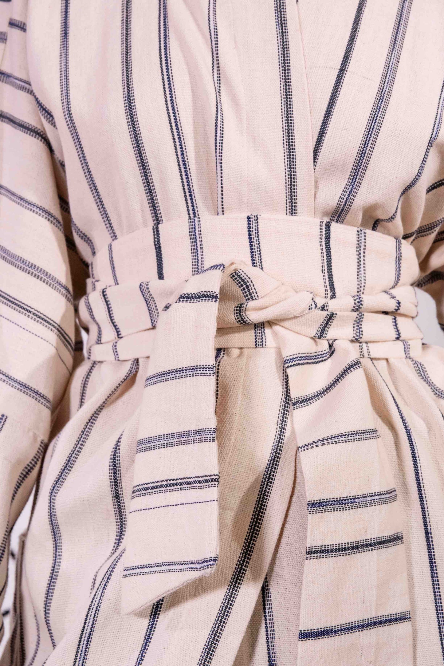 Model is wearing a midi-length, handwoven and hand-printed Benin cotton kimono by Atelier V.O, featuring traditional Dogon and Nagô-inspired patterns, a straight cut, drawstring closure, and wooden buttons.