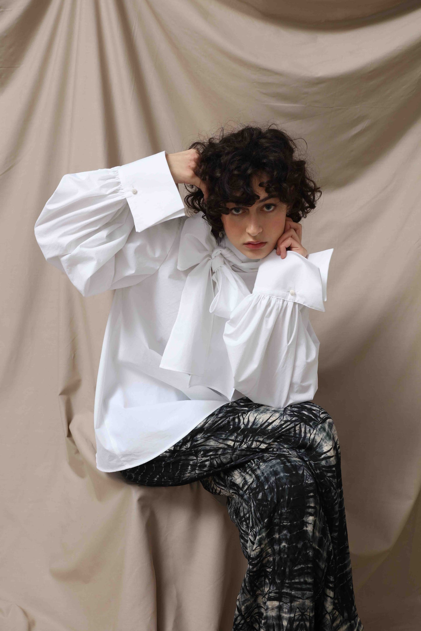 Regula Blouse by House of Woven Stories, handcrafted in Italy from 100% organic cotton poplin. Features adjustable long bow, minimalist tailoring, and sustainable design inspired by global craftsmanship.