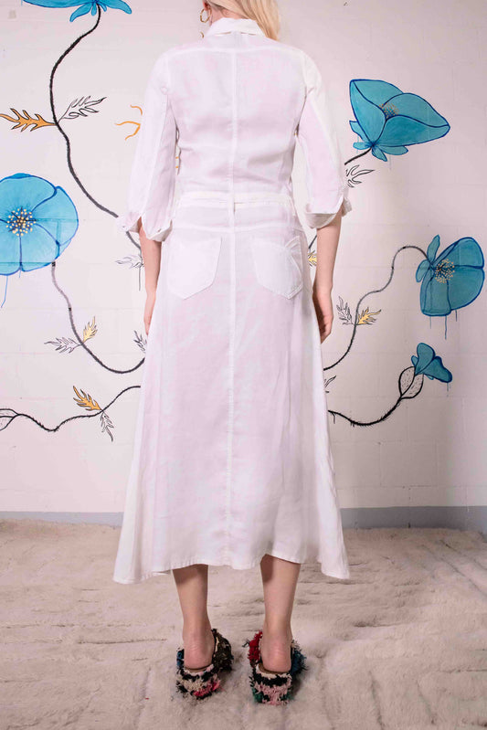 Model is wearing the Blanc Cassé Trompe L’Oeil dress by Happy Haus, made from a soft hemp and silk blend with a classic collar, bronze buttons, 7/8-length pleated sleeves, and floral patch pockets including peacock embroidery.
