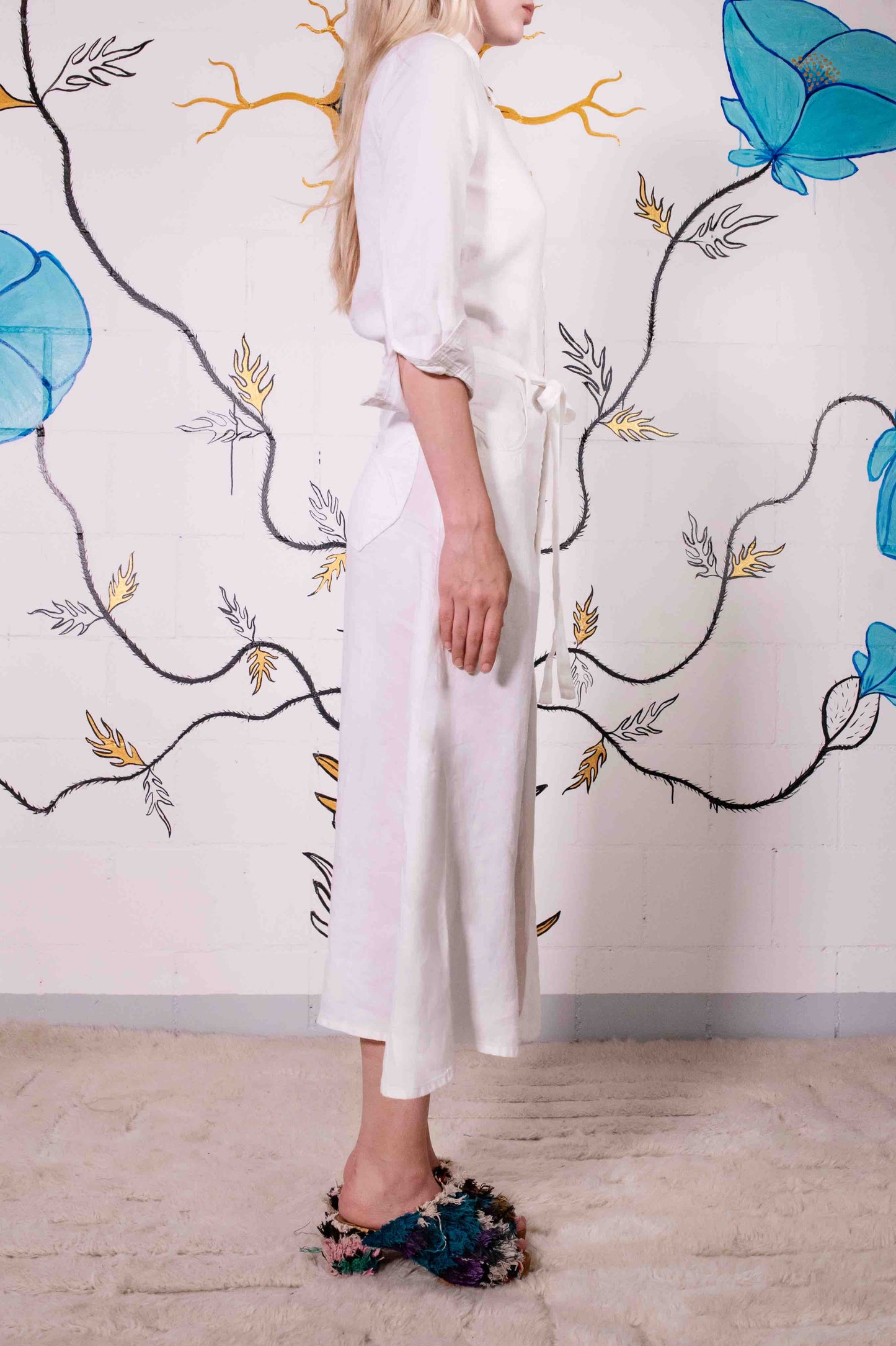 Model is wearing the Blanc Cassé Trompe L’Oeil dress by Happy Haus, made from a soft hemp and silk blend with a classic collar, bronze buttons, 7/8-length pleated sleeves, and floral patch pockets including peacock embroidery.