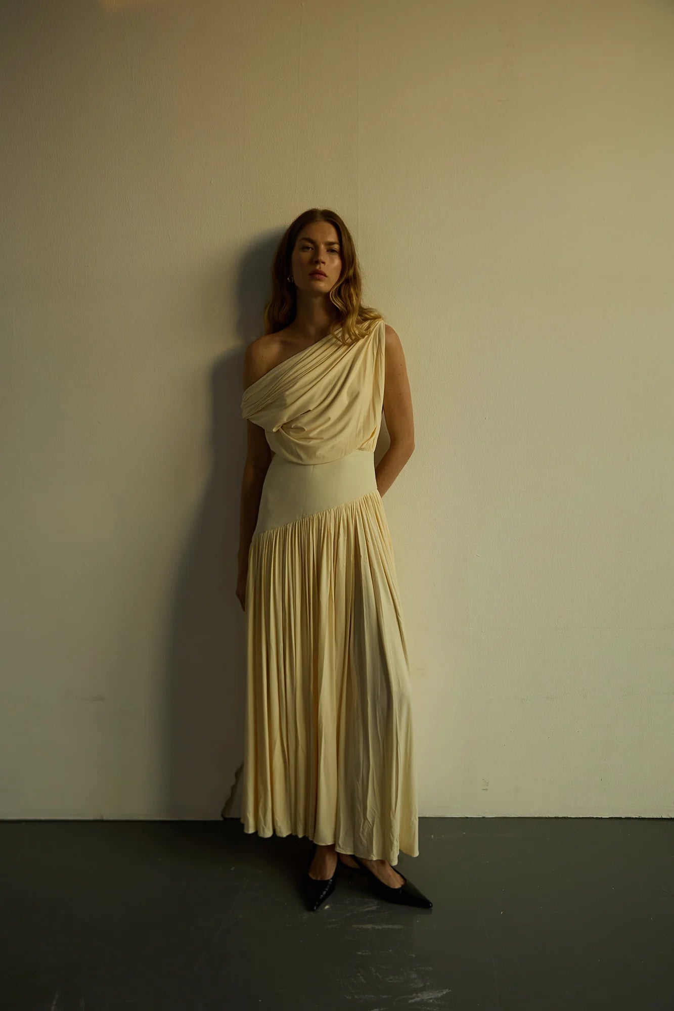 Model wearing a cream Gwen Off Shoulder Drape Dress by Abadia from the House of Nomad Stories collection, featuring a stretch jersey fabric, asymmetrical curved waistband, and a sculpted elegant maxi silhouette.