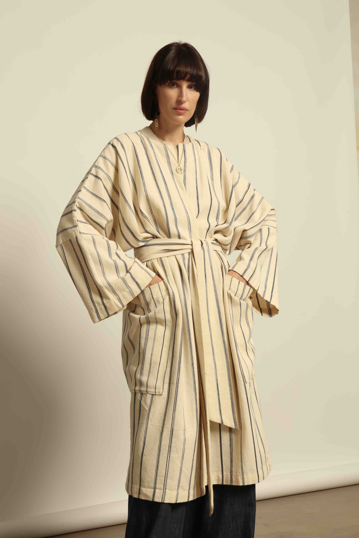 Model is wearing the Maxi Handwoven and Hand-Printed Benin Cotton Kimono ‘The Ones Who Laugh’ by Atelier V.O, crafted from 100% handwoven cotton with Dogon Mali-inspired print, straight cut, drawstring, wooden button fastening, and matching belt.
