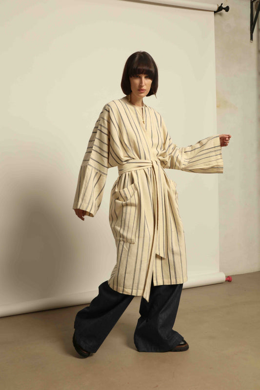 Model is wearing the Maxi Handwoven and Hand-Printed Benin Cotton Kimono ‘The Ones Who Laugh’ by Atelier V.O, crafted from 100% handwoven cotton with Dogon Mali-inspired print, straight cut, drawstring, wooden button fastening, and matching belt.