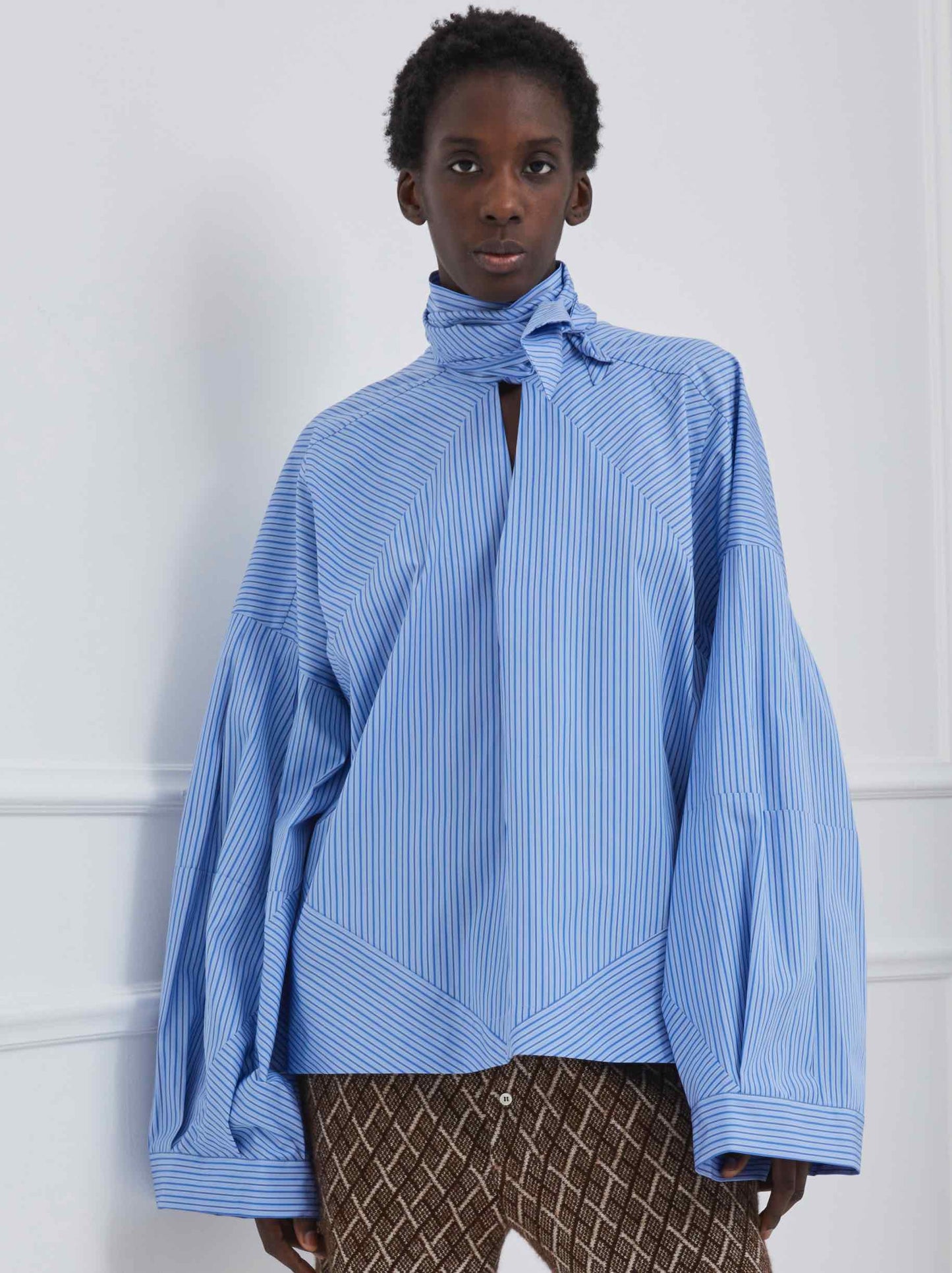 Model is wearing the Clo Blue Stripes Shirt by Maison Jejia, part of the House of Woven Stories selection, featuring puffed sleeves, wrap-style design, and subtle blue stripes.