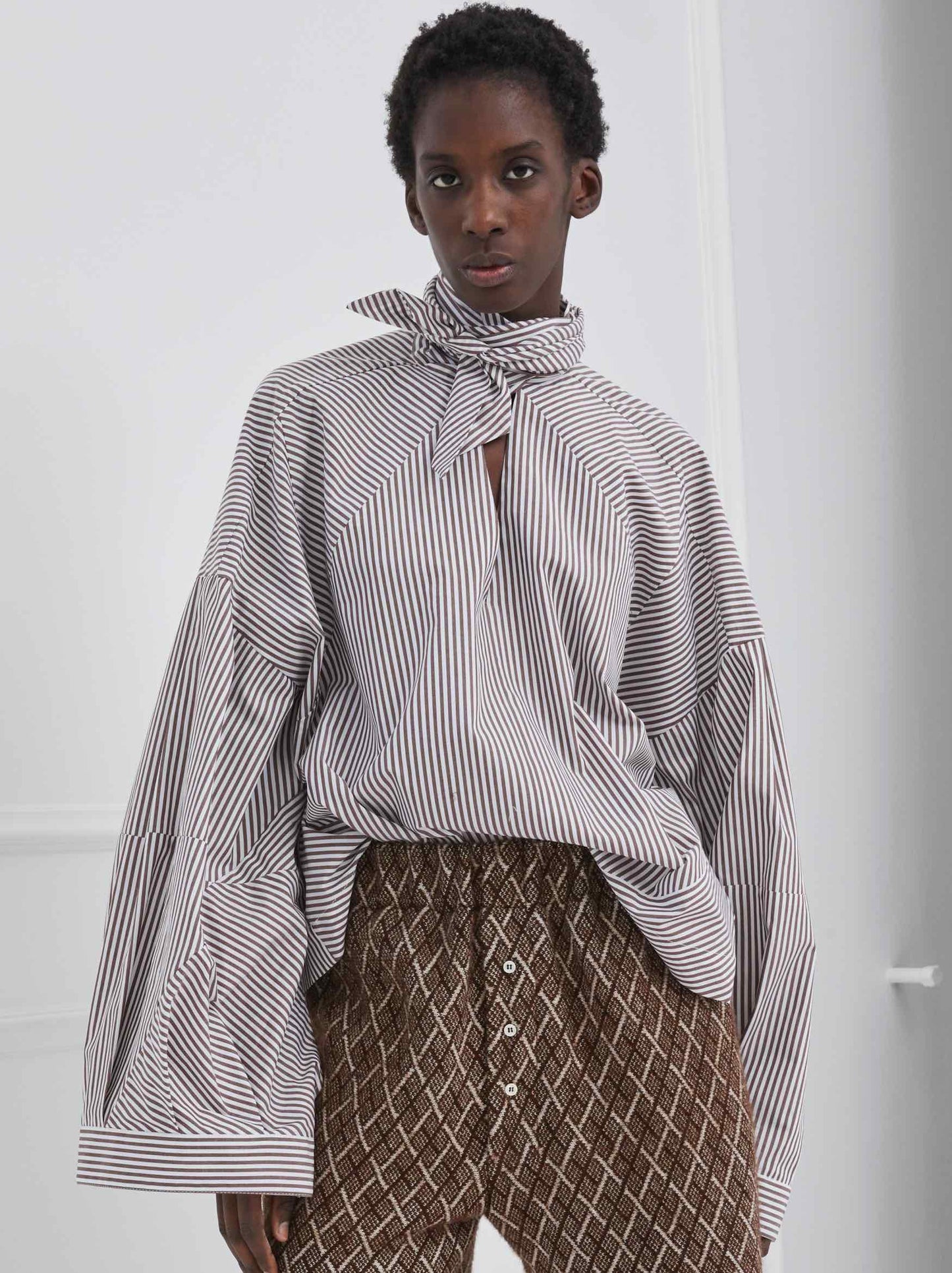 Model is wearing the Clo Brown and White Cotton Stripes Shirt by Maison Jejia, part of the House of Woven Stories selection, featuring puffed sleeves, wrap-style design, and warm brown stripes.