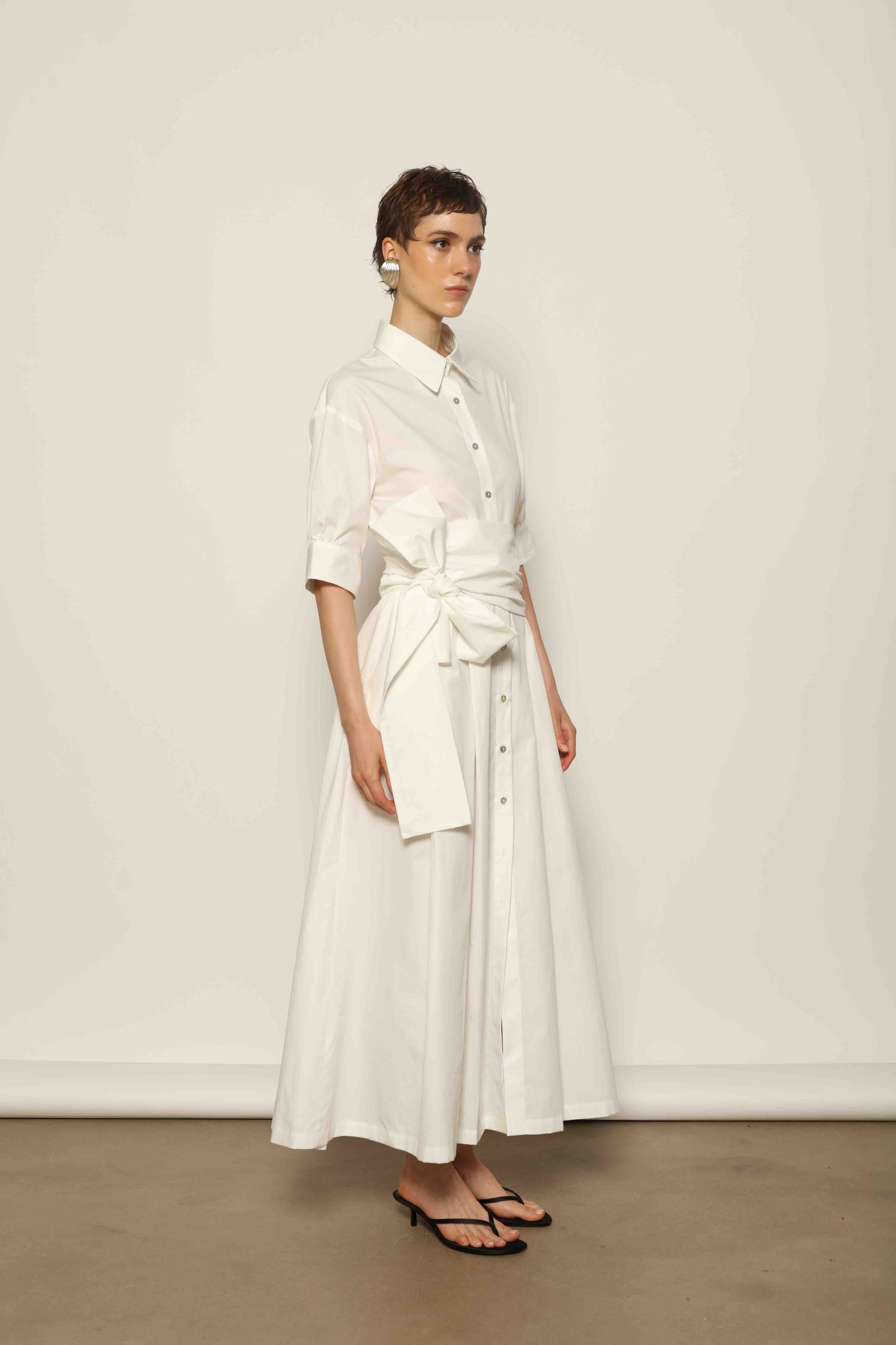 Model wearing the Emily Organic White Cotton Dress by House of Woven Stories, featuring 100% organic cotton fabric, classic collar, button-down front, and adjustable belt for a clean, elegant silhouette.