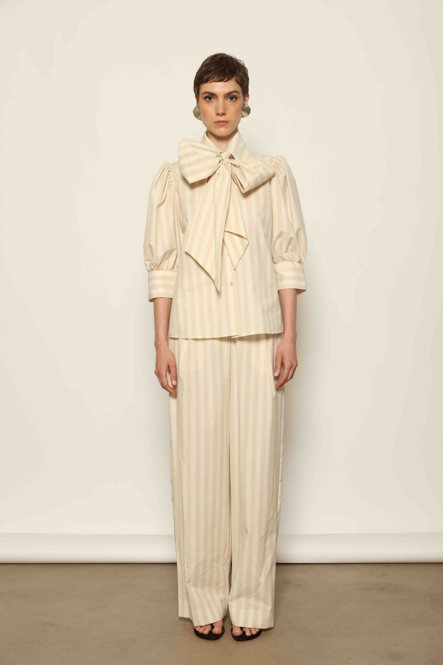 Model wearing the Gabriella Sand White Striped Trousers by House of Woven Stories, featuring a high-rise waist, straight-leg tailored fit, and soft sand and white stripe pattern in a premium cotton, merino wool, and cashmere blend.