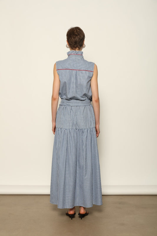 Model wearing the Karin Vichy Blue Dress by House of Woven Stories, featuring 100% organic cotton fabric, blue gingham pattern, sleeveless silhouette, detachable belt, and ruched collar with red piping detail.