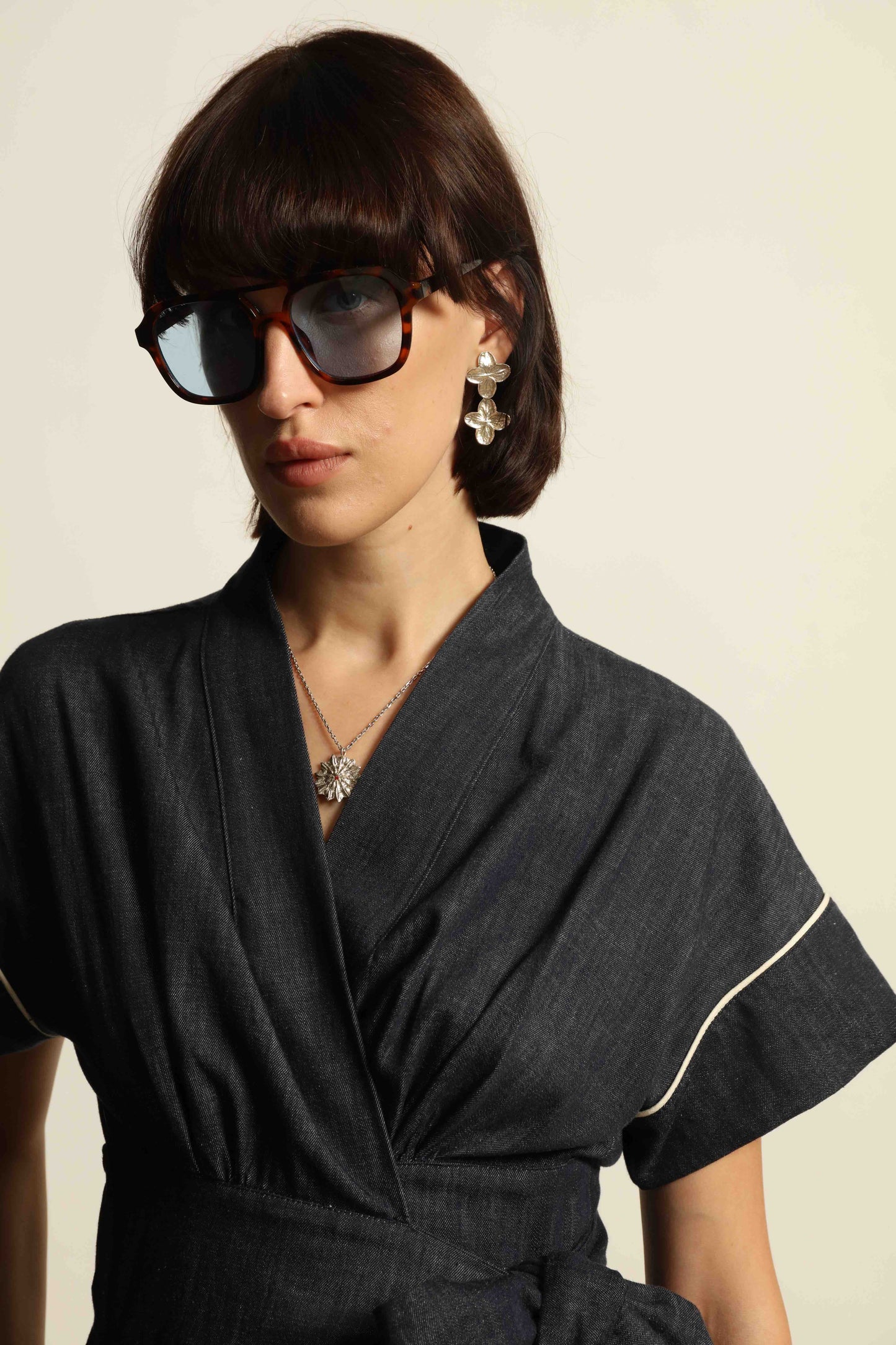 Model is wearing the Saint Remy wrap dress by House of Woven Stories in 100% cotton, featuring a denim-blue color with white pinstripes, drop shoulders, short sleeves, deep V-neckline, and self-tie belt, handmade in Italy.