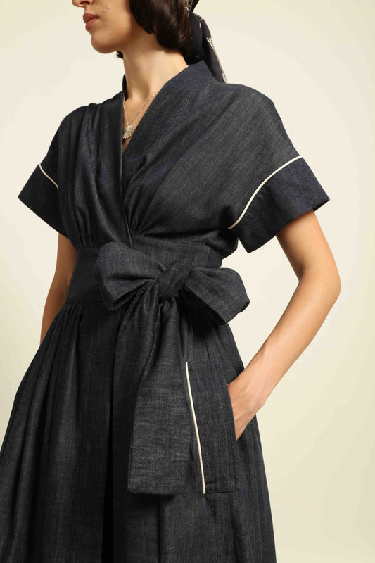 Model is wearing the Saint Remy wrap dress by House of Woven Stories in 100% cotton, featuring a denim-blue color with white pinstripes, drop shoulders, short sleeves, deep V-neckline, and self-tie belt, handmade in Italy.