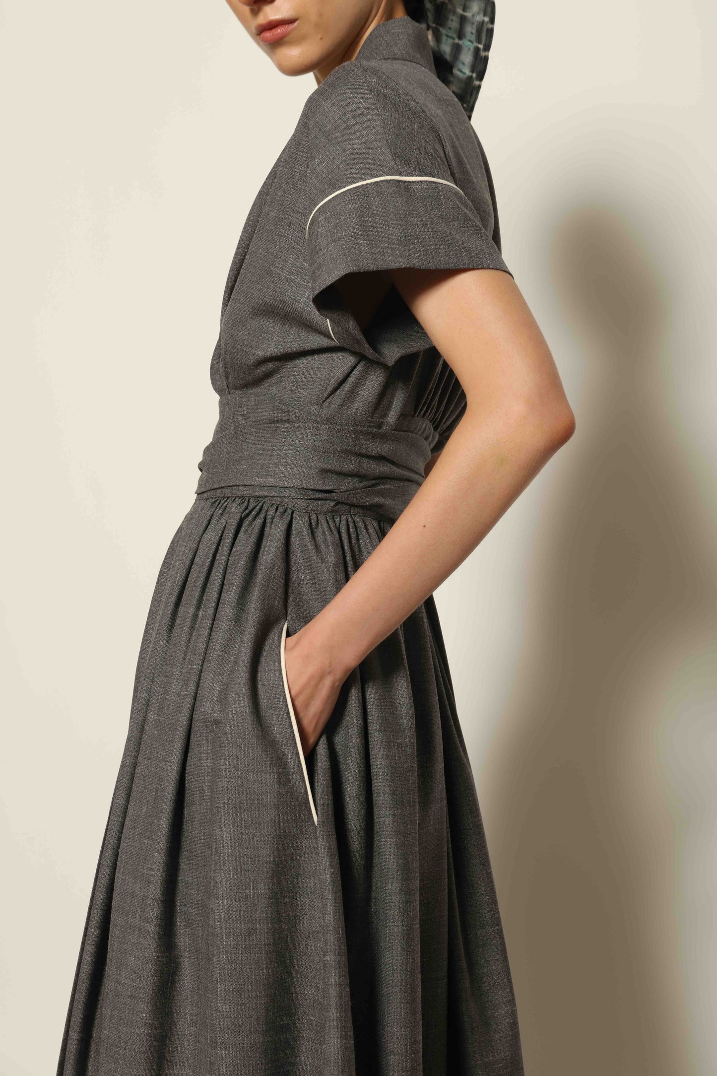 Model wearing the Saint Remy Grey Off White Midi Dress by House of Woven Stories, featuring a wrap silhouette, adjustable belt, half sleeves, and subtle white piping detail in a fluid elegant fabric.
