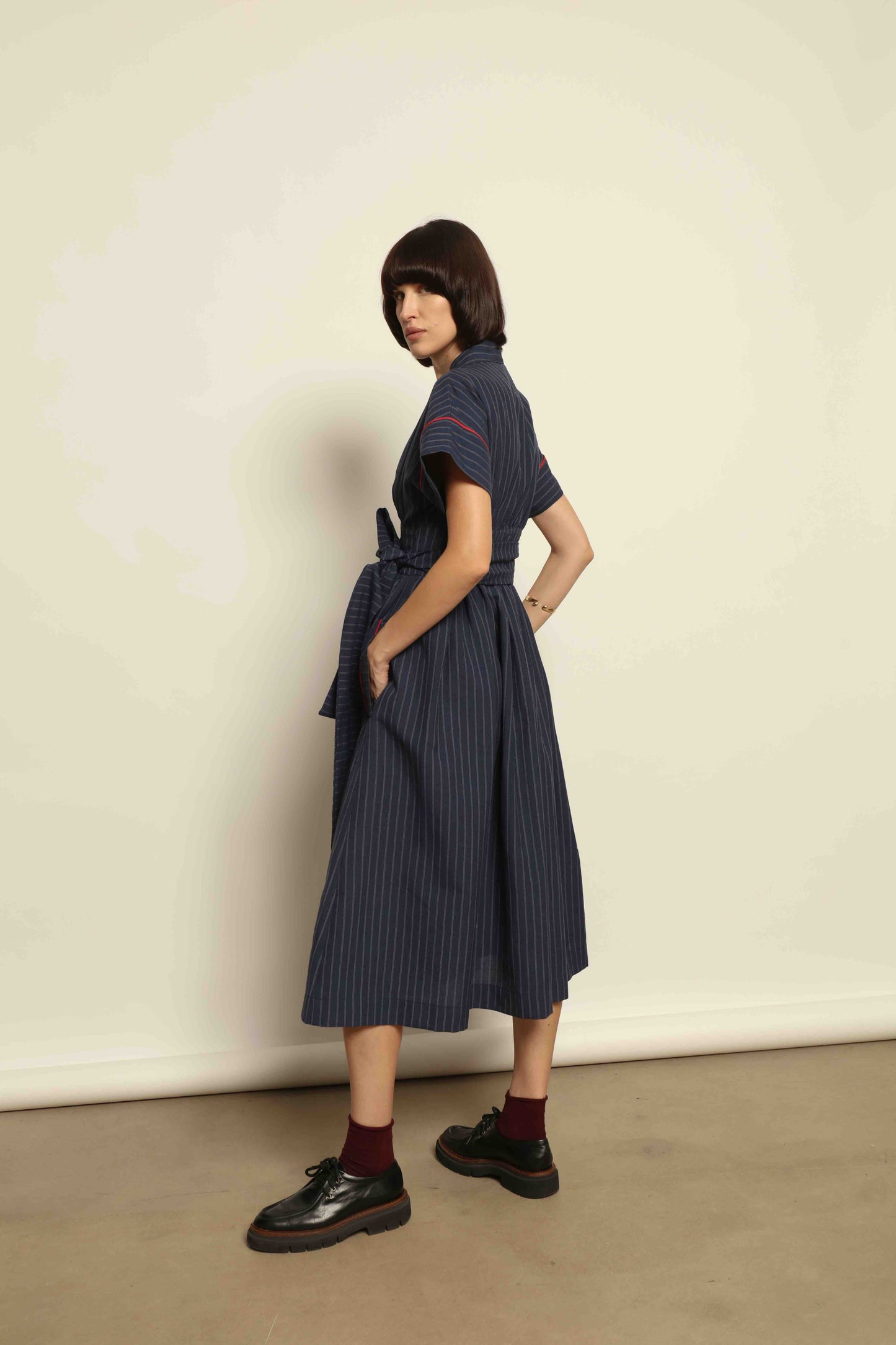 Model is wearing the Saint Remy wrap dress by House of Woven Stories in a blue and white pinstripe, made from a breathable wool-linen blend, featuring drop shoulders, short sleeves, deep V-neckline, and a matching fabric belt, handmade in Italy.