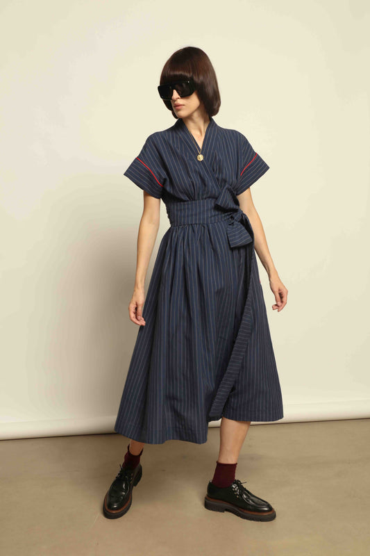 Model is wearing the Saint Remy wrap dress by House of Woven Stories in a blue and white pinstripe, made from a breathable wool-linen blend, featuring drop shoulders, short sleeves, deep V-neckline, and a matching fabric belt, handmade in Italy.