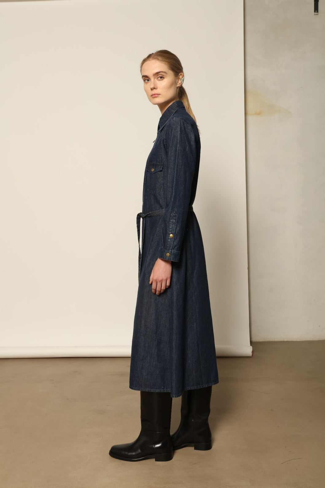 Model is wearing La Robe Star in Bleu D’été by Happy Haus, a denim shirt dress with relaxed fit, chest pockets, matching belt, and golden button detail, combining timeless style with versatile year-round wear.