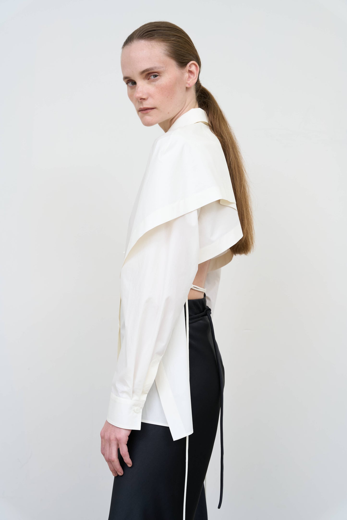 Model is wearing a fitted white cotton shirt by Litkovska with capelet overlay and open-back detail, architectural designer top available at House of Woven Stories
