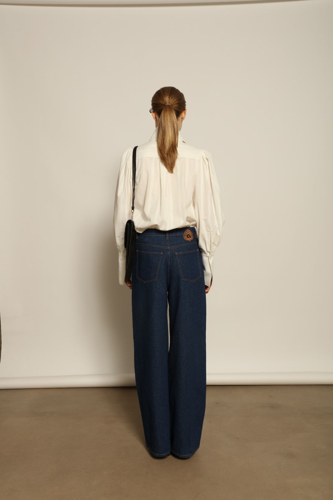 Model is wearing Le Pantalon Peacock in Brut Vintage by Happy Haus, high-waisted wide-leg cotton trousers with double-stitched back pockets and delicate embroidered peacock detail, combining timeless elegance with relaxed sophistication.