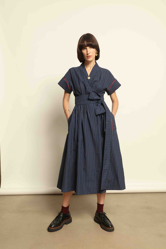 Model is wearing the Saint Remy wrap dress by House of Woven Stories in a blue and white pinstripe, made from a breathable wool-linen blend, featuring drop shoulders, short sleeves, deep V-neckline, and a matching fabric belt, handmade in Italy.