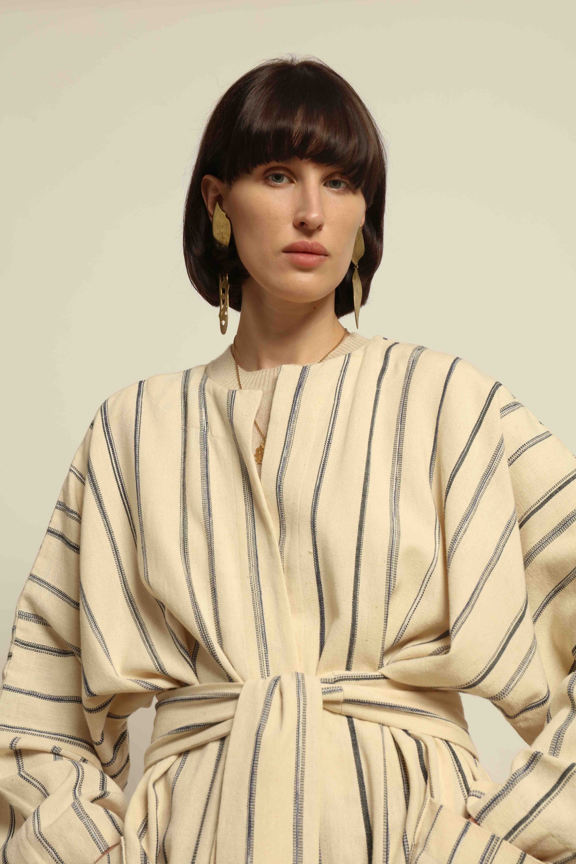 Model is wearing the Maxi Handwoven and Hand-Printed Benin Cotton Kimono ‘The Ones Who Laugh’ by Atelier V.O, crafted from 100% handwoven cotton with Dogon Mali-inspired print, straight cut, drawstring, wooden button fastening, and matching belt.