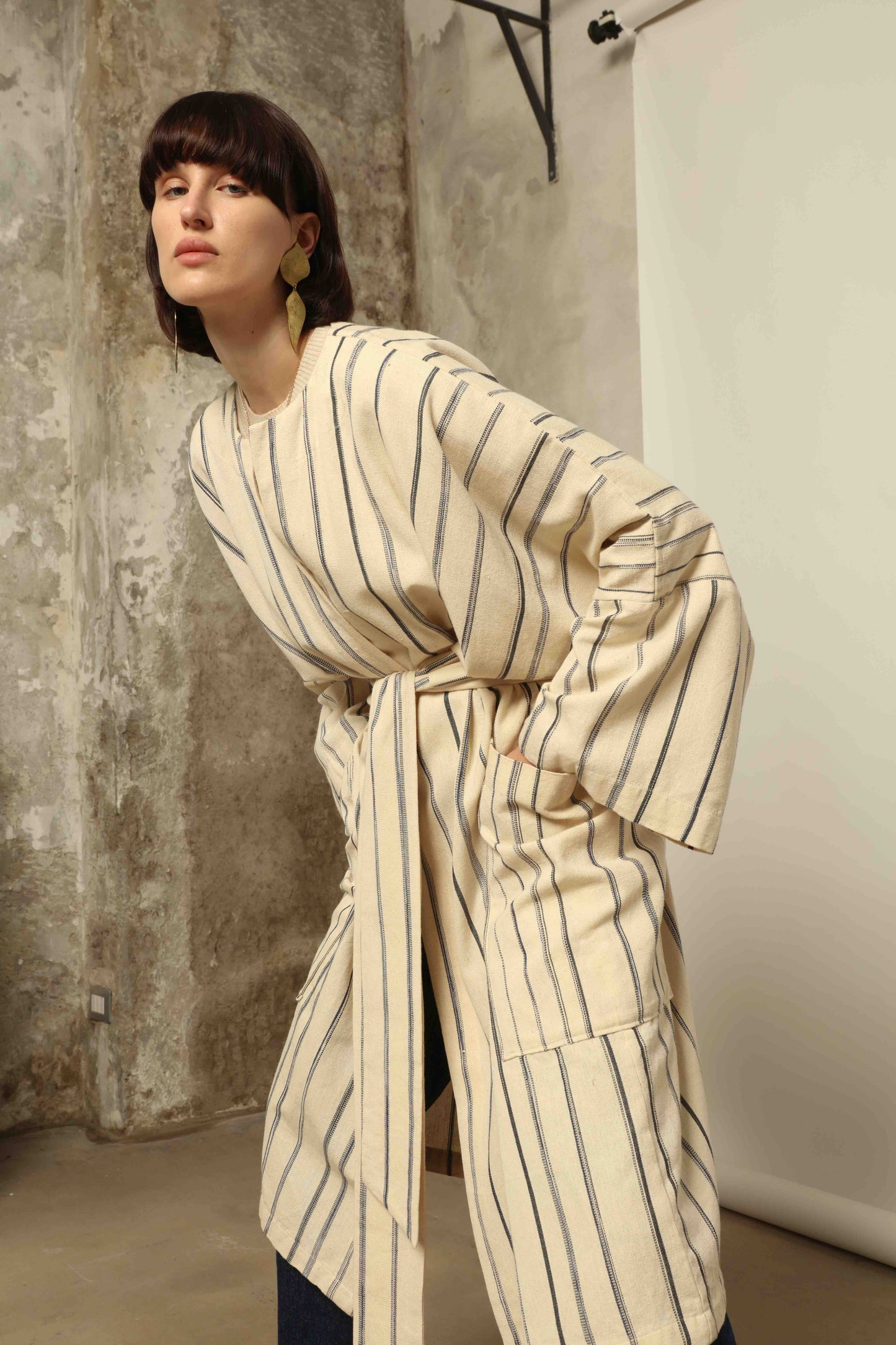 Model is wearing the Maxi Handwoven and Hand-Printed Benin Cotton Kimono ‘The Ones Who Laugh’ by Atelier V.O, crafted from 100% handwoven cotton with Dogon Mali-inspired print, straight cut, drawstring, wooden button fastening, and matching belt.