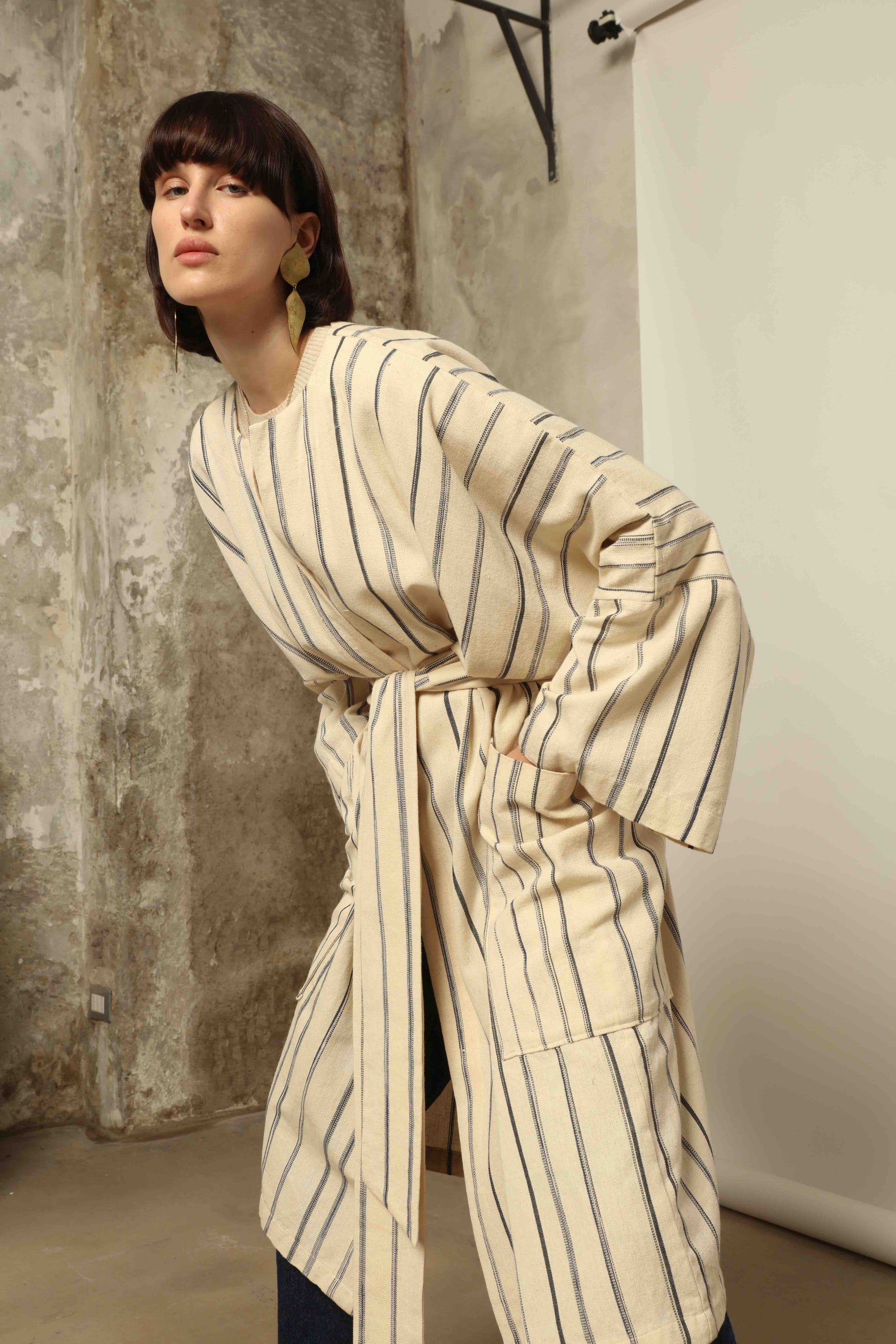 Model is wearing the Maxi Handwoven and Hand-Printed Benin Cotton Kimono ‘The Ones Who Laugh’ by Atelier V.O, crafted from 100% handwoven cotton with Dogon Mali-inspired print, straight cut, drawstring, wooden button fastening, and matching belt.