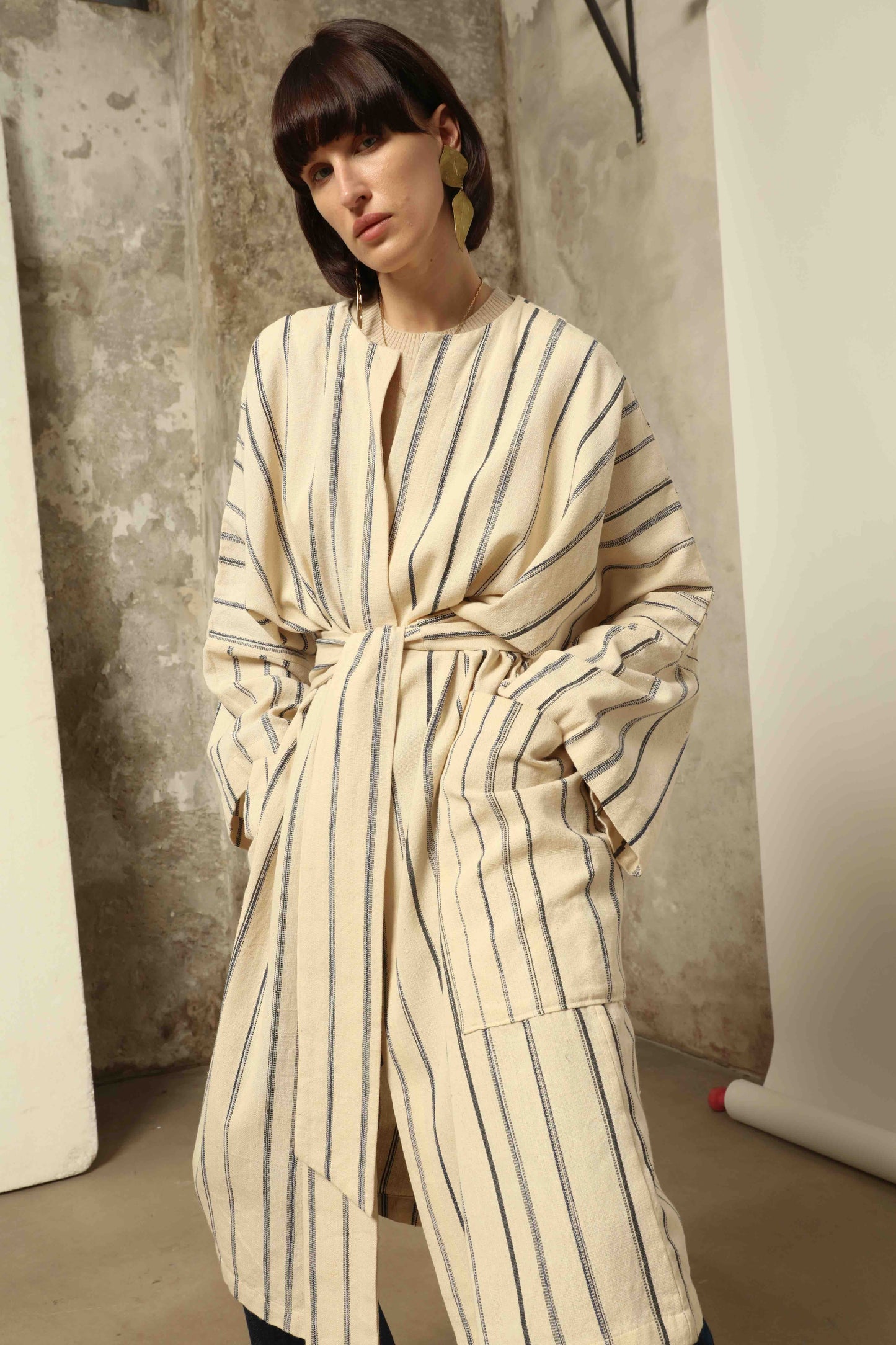 Model is wearing the Maxi Handwoven and Hand-Printed Benin Cotton Kimono ‘The Ones Who Laugh’ by Atelier V.O, crafted from 100% handwoven cotton with Dogon Mali-inspired print, straight cut, drawstring, wooden button fastening, and matching belt.