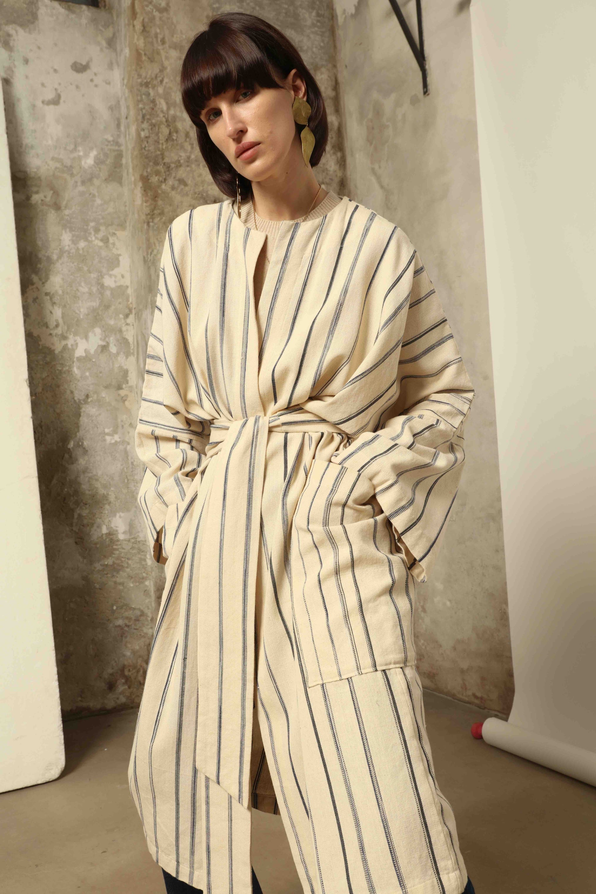 Model is wearing the Maxi Handwoven and Hand-Printed Benin Cotton Kimono ‘The Ones Who Laugh’ by Atelier V.O, crafted from 100% handwoven cotton with Dogon Mali-inspired print, straight cut, drawstring, wooden button fastening, and matching belt.