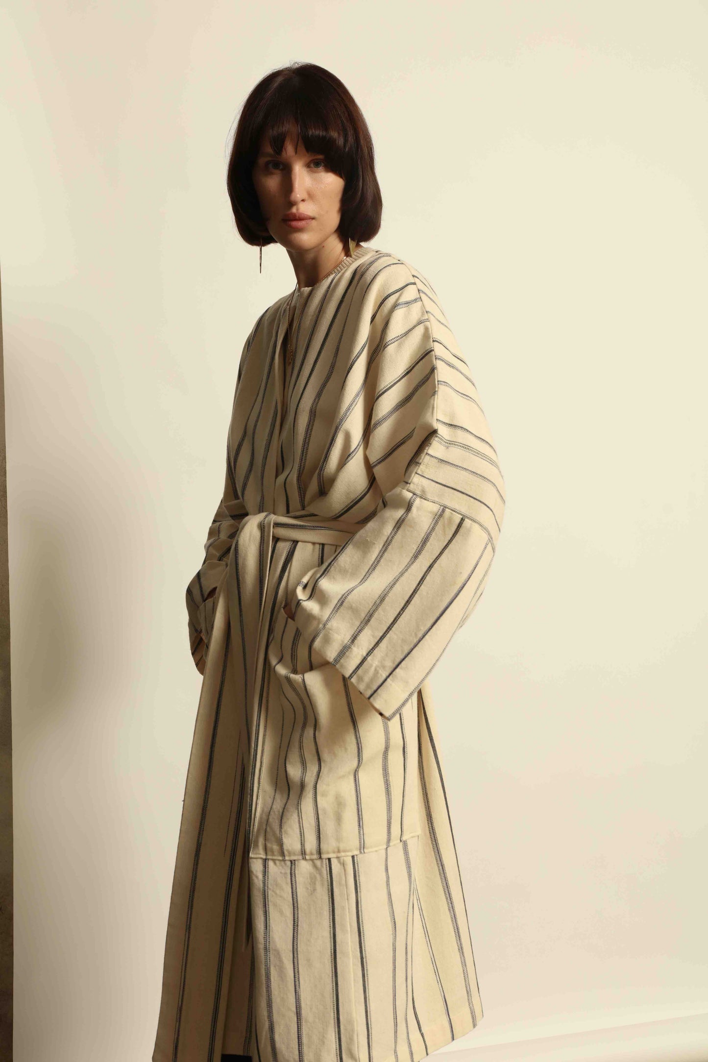 Model is wearing the Maxi Handwoven and Hand-Printed Benin Cotton Kimono ‘The Ones Who Laugh’ by Atelier V.O, crafted from 100% handwoven cotton with Dogon Mali-inspired print, straight cut, drawstring, wooden button fastening, and matching belt.