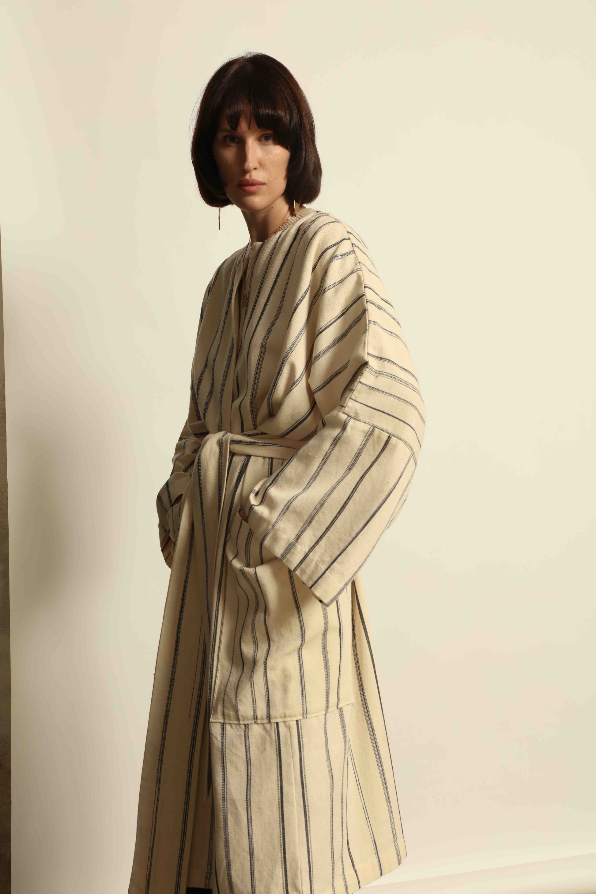 Model is wearing the Maxi Handwoven and Hand-Printed Benin Cotton Kimono ‘The Ones Who Laugh’ by Atelier V.O, crafted from 100% handwoven cotton with Dogon Mali-inspired print, straight cut, drawstring, wooden button fastening, and matching belt.