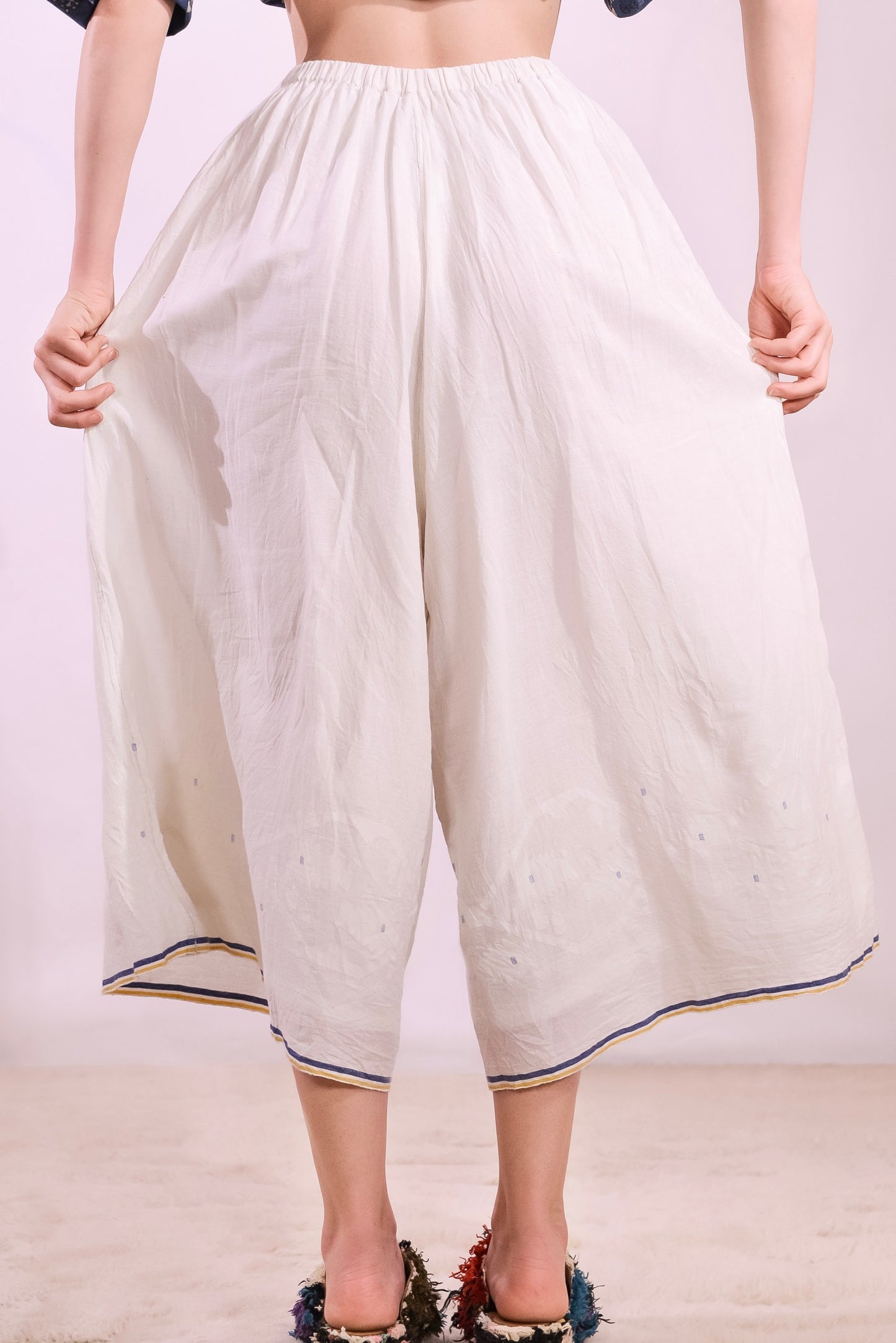 Model is wearing the Amber 59 handloomed pyjama trousers by Injiri — wide-leg organic cotton pants in crisp white with cobalt and yellow jamdani weaving along the hem, featuring an elasticated back waistband and side pockets.