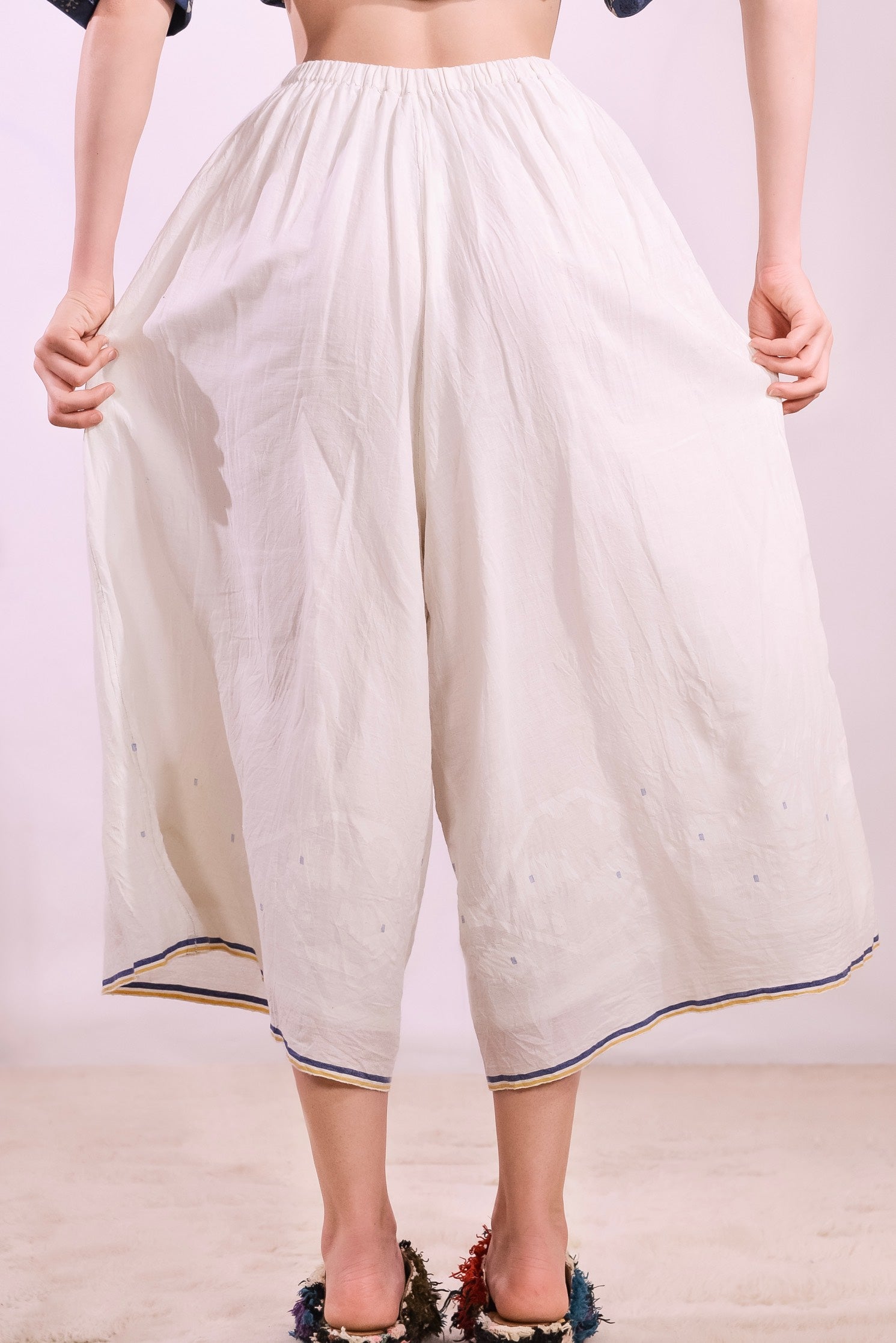 Model is wearing the Amber 59 handloomed pyjama trousers by Injiri — wide-leg organic cotton pants in crisp white with cobalt and yellow jamdani weaving along the hem, featuring an elasticated back waistband and side pockets.