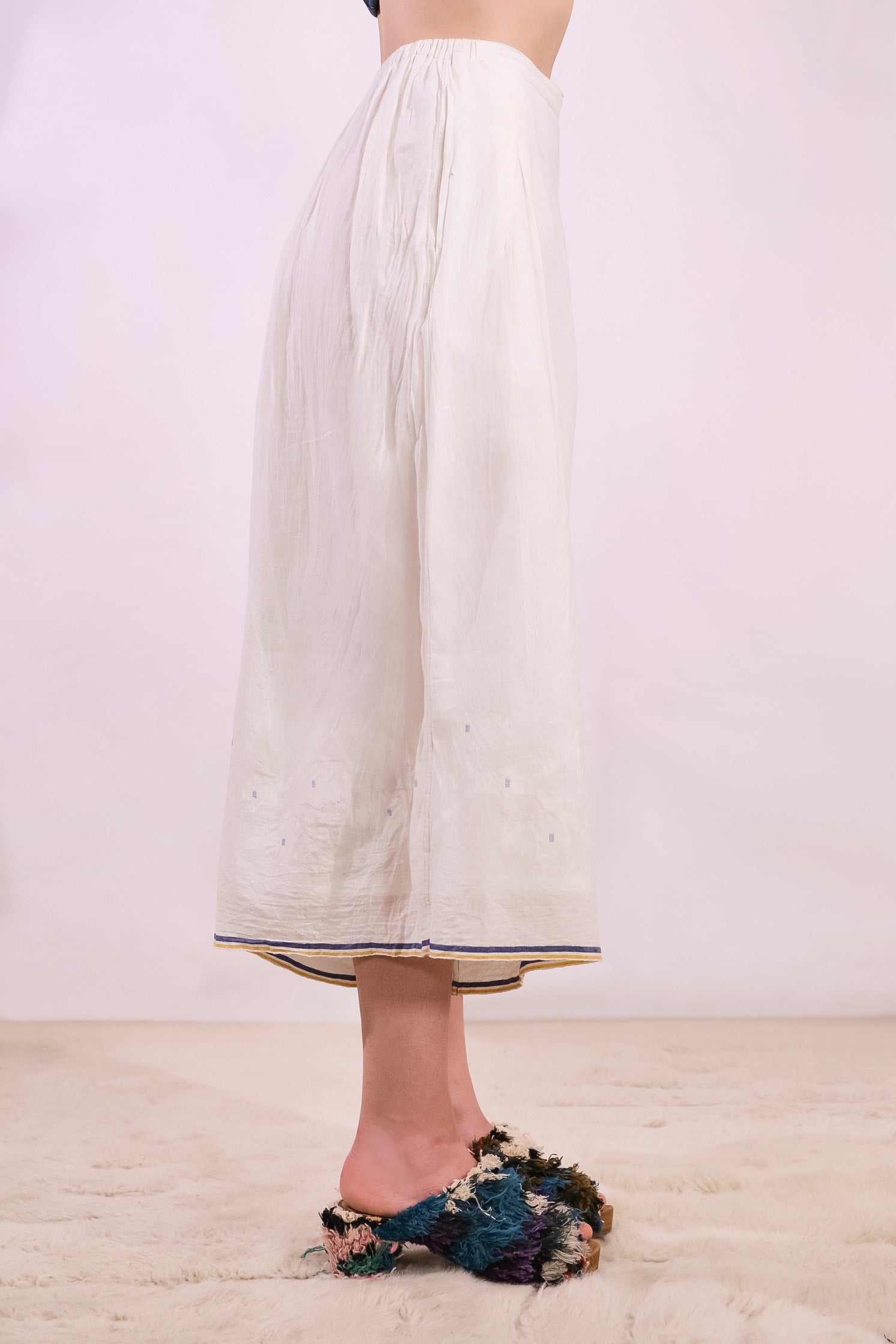 Model is wearing the Amber 59 handloomed pyjama trousers by Injiri — wide-leg organic cotton pants in crisp white with cobalt and yellow jamdani weaving along the hem, featuring an elasticated back waistband and side pockets.