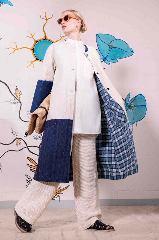 Model is wearing a reversible quilted cotton coat by Injiri, featuring a tie-dye design in off-white and deep indigo, with a split rounded collar, contrast buttons, and blue madras check lining.