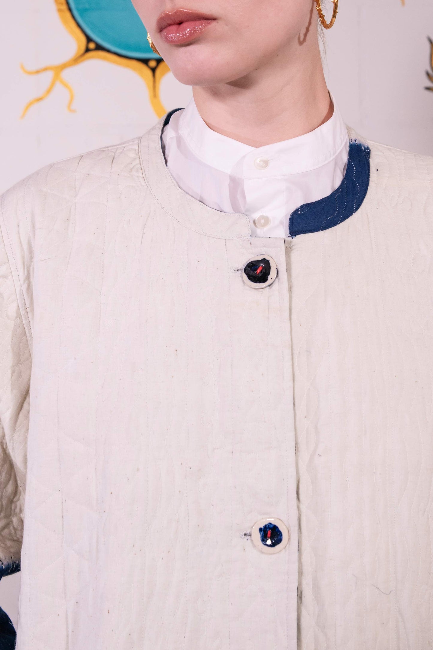 Model is wearing a reversible quilted cotton coat by Injiri, featuring a tie-dye design in off-white and deep indigo, with a split rounded collar, contrast buttons, and blue madras check lining.