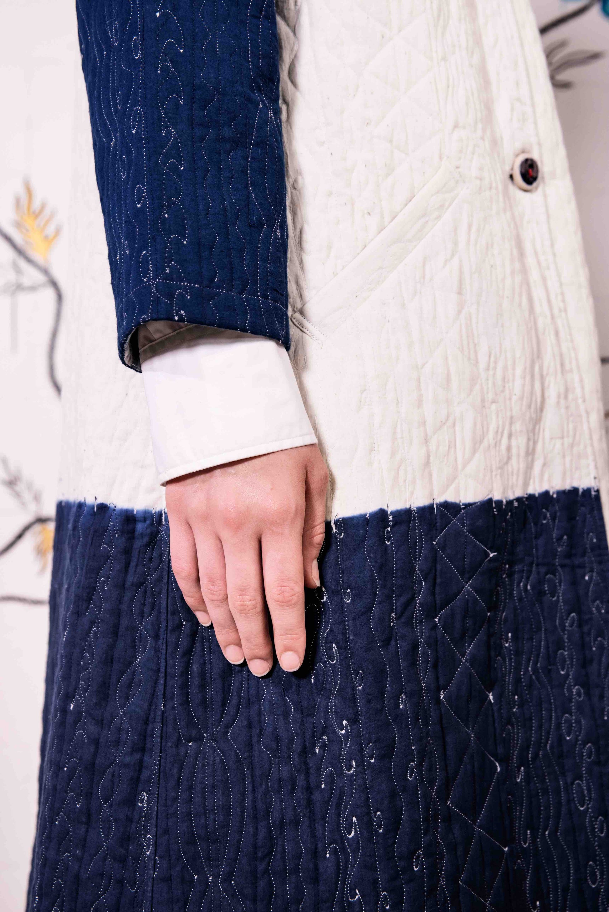 Model is wearing a reversible quilted cotton coat by Injiri, featuring a tie-dye design in off-white and deep indigo, with a split rounded collar, contrast buttons, and blue madras check lining.