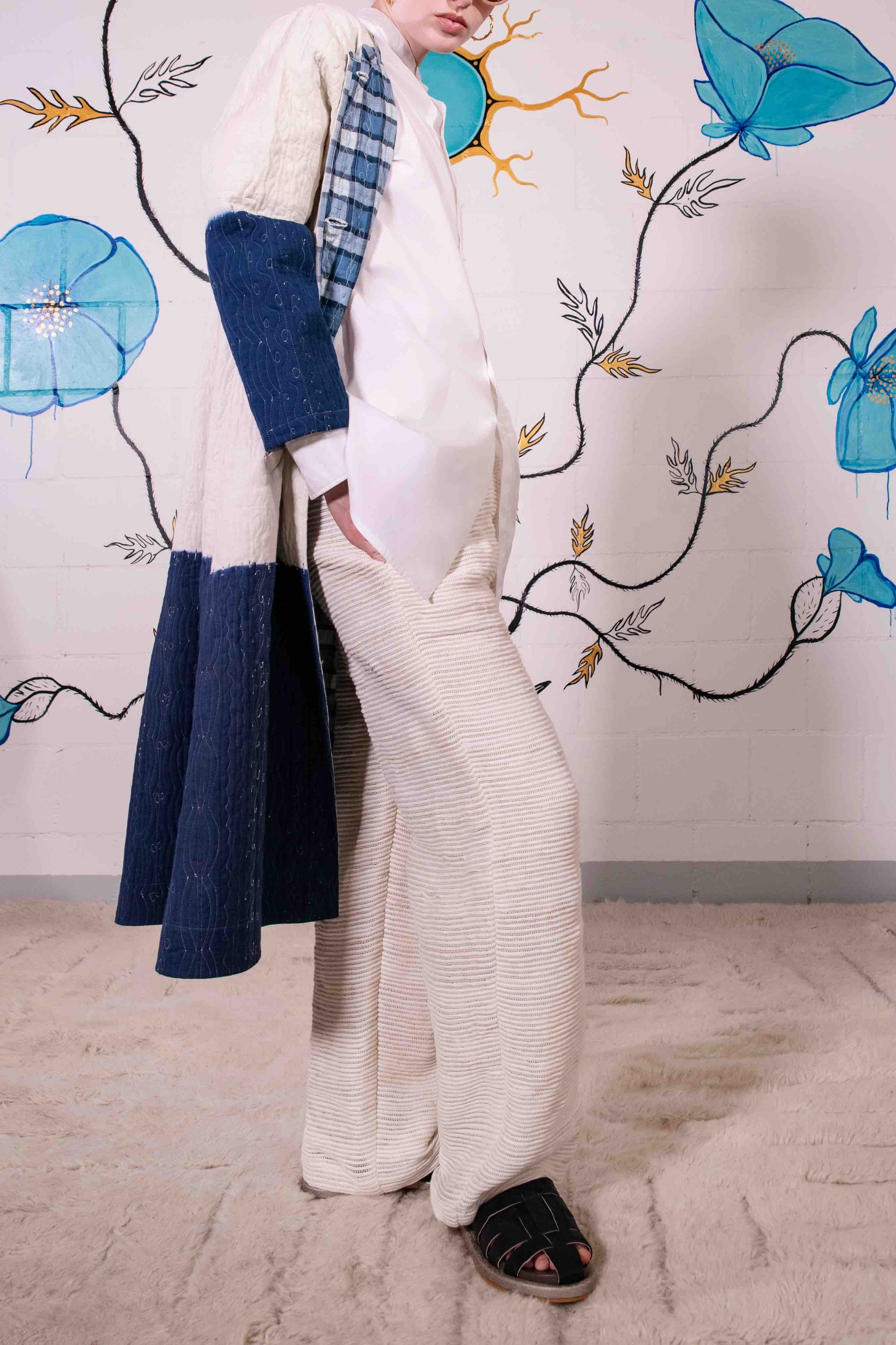 Model is wearing a reversible quilted cotton coat by Injiri, featuring a tie-dye design in off-white and deep indigo, with a split rounded collar, contrast buttons, and blue madras check lining.