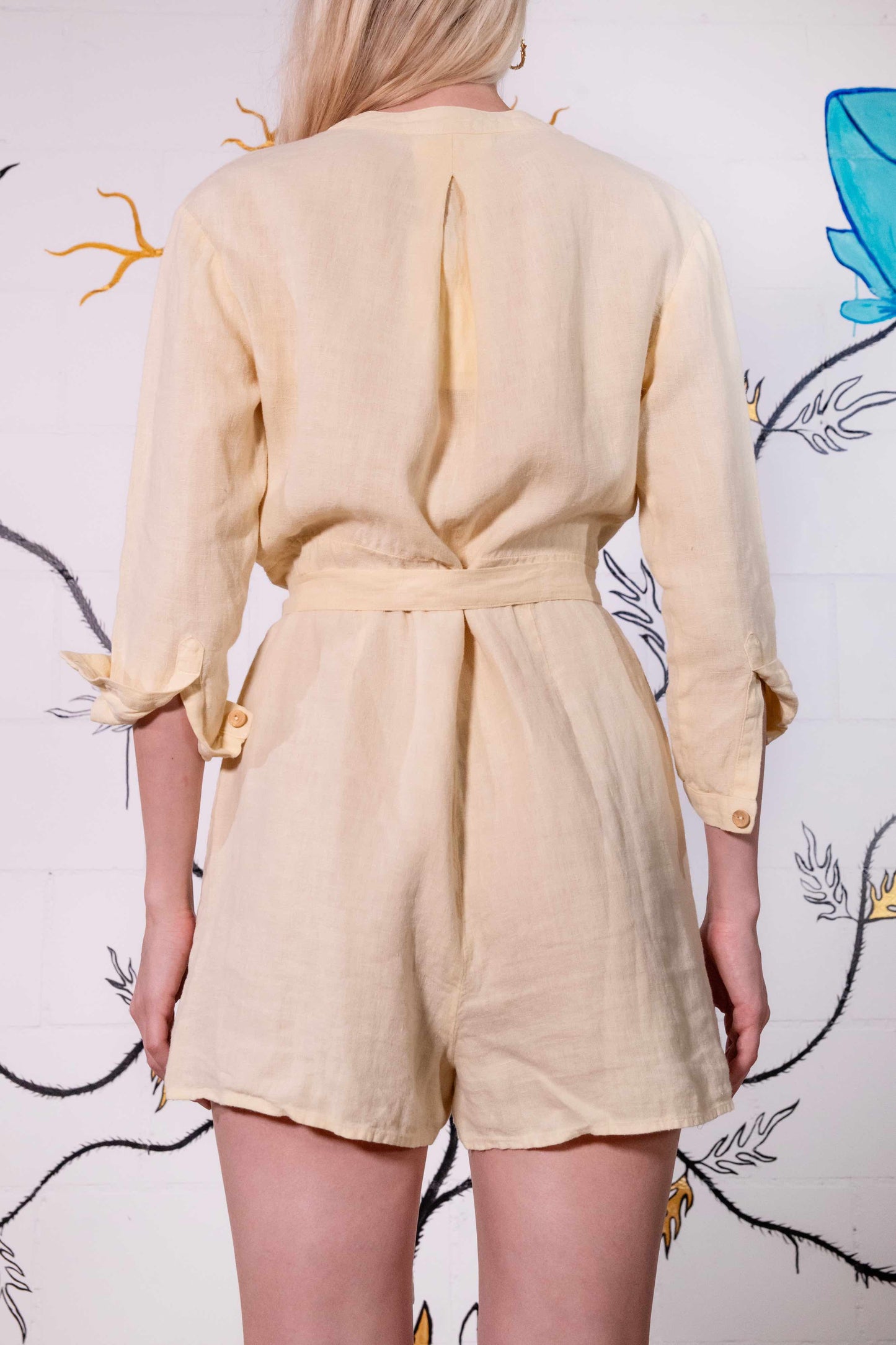 Model is wearing a banana yellow short linen jumpsuit by Happy Haus, featuring a relaxed fit, oversized chest and side pockets, wooden buttons, and a matching fabric belt.