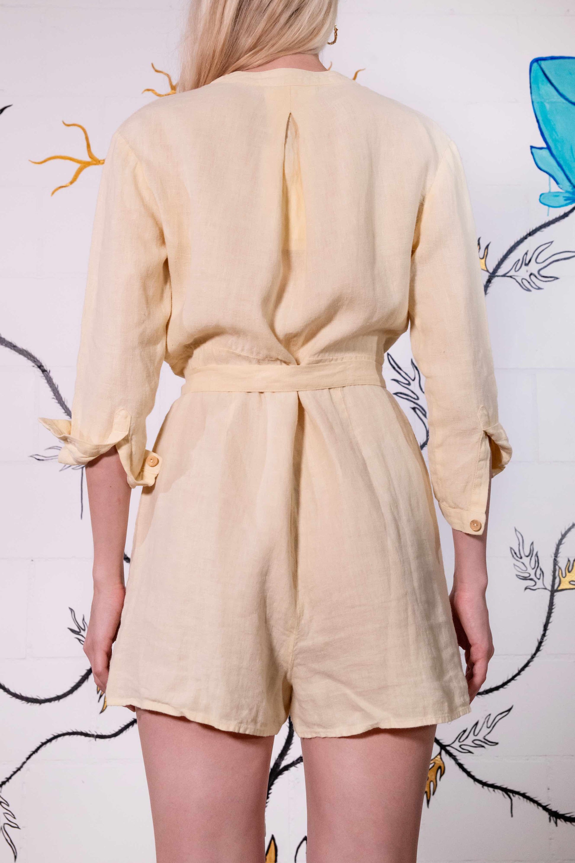 Model is wearing a banana yellow short linen jumpsuit by Happy Haus, featuring a relaxed fit, oversized chest and side pockets, wooden buttons, and a matching fabric belt.