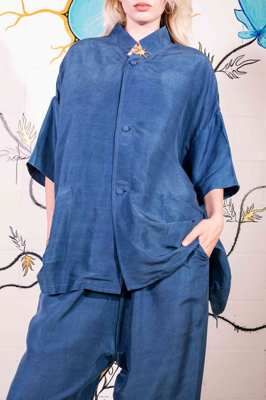 Model is wearing a short-sleeve indigo shirt made from 100% silk, featuring a relaxed fit, dropped shoulders, front button closure, and large patch pockets. Hand-dyed using botanical pigments by A Sacred Wrap.