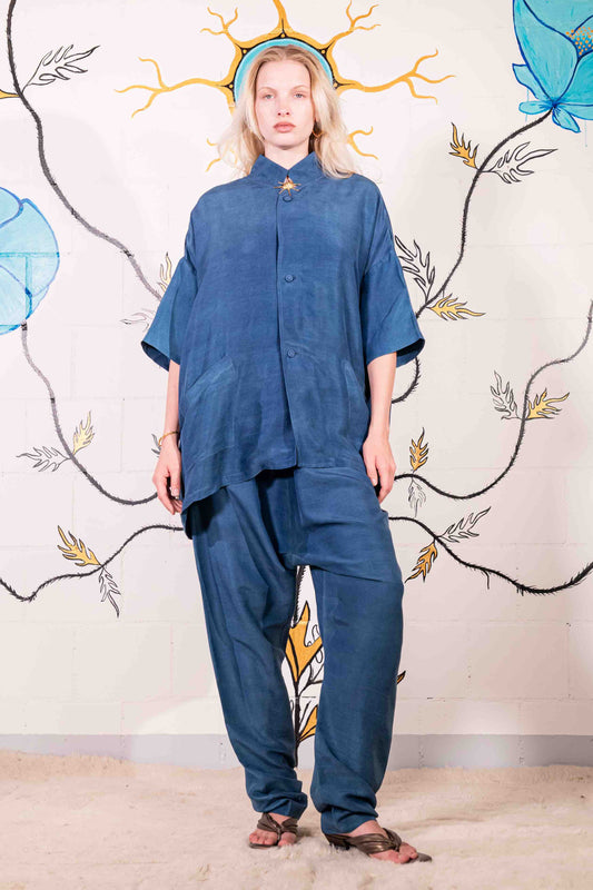 Model is wearing a short-sleeve indigo shirt made from 100% silk, featuring a relaxed fit, dropped shoulders, front button closure, and large patch pockets. Hand-dyed using botanical pigments by A Sacred Wrap.