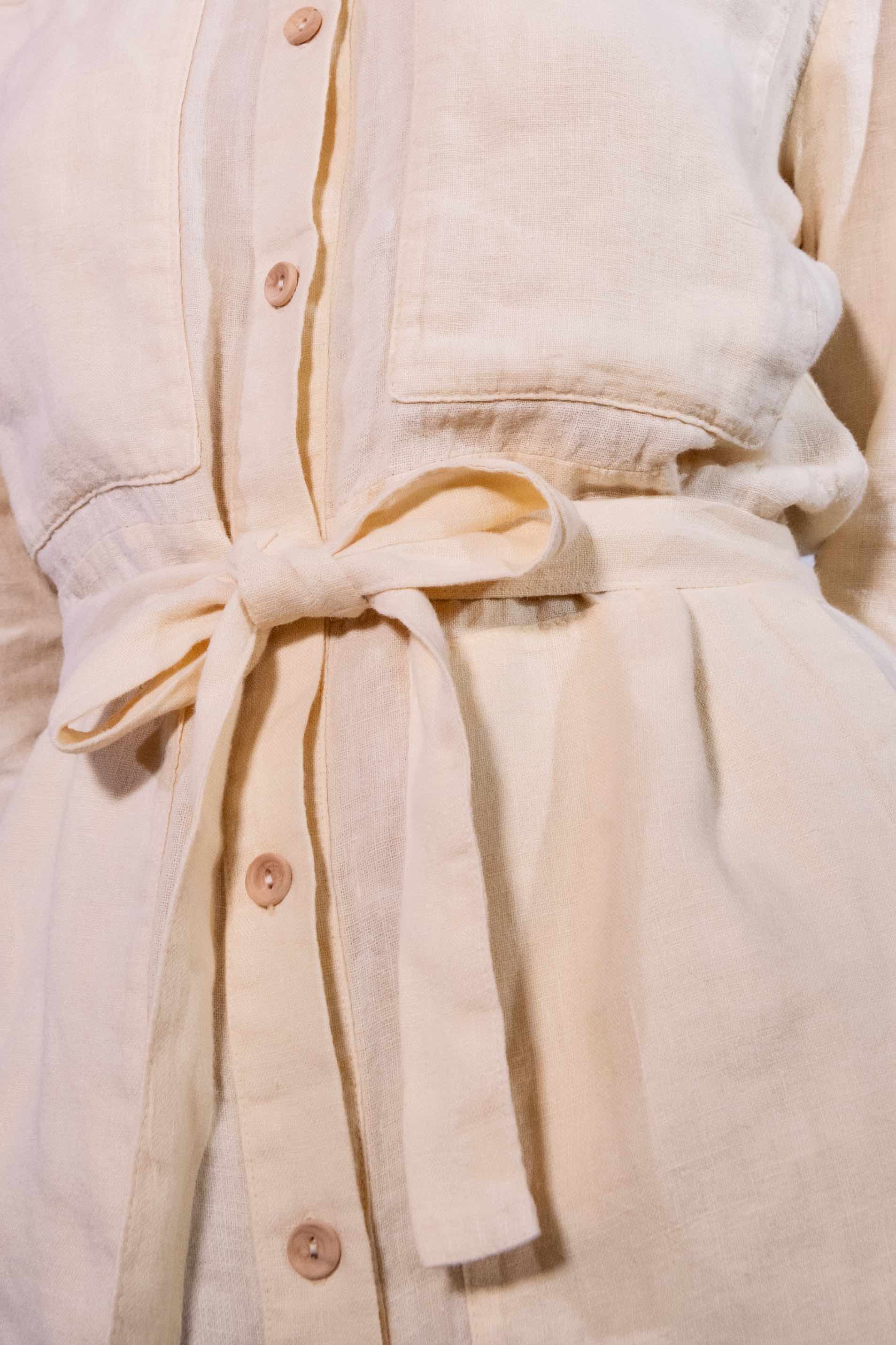 Model is wearing a banana yellow short linen jumpsuit by Happy Haus, featuring a relaxed fit, oversized chest and side pockets, wooden buttons, and a matching fabric belt.