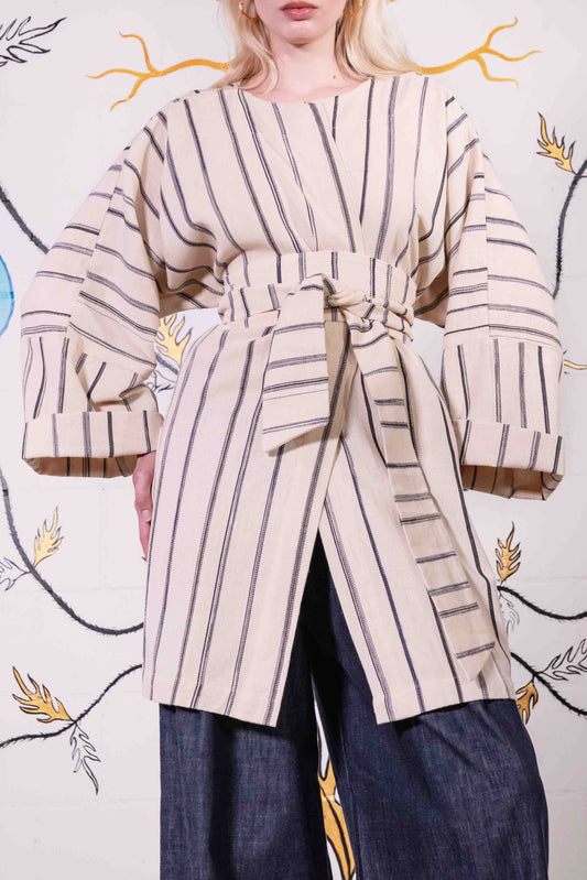 Model is wearing a midi-length, handwoven and hand-printed Benin cotton kimono by Atelier V.O, featuring traditional Dogon and Nagô-inspired patterns, a straight cut, drawstring closure, and wooden buttons.