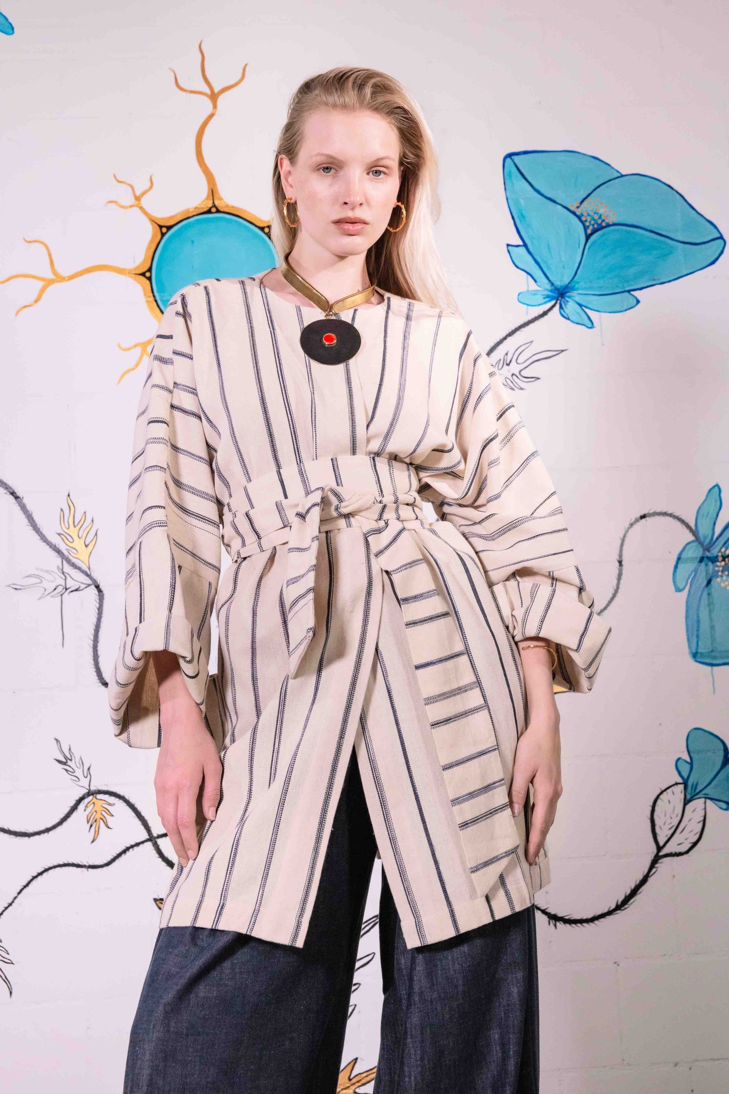 Model is wearing a midi-length, handwoven and hand-printed Benin cotton kimono by Atelier V.O, featuring traditional Dogon and Nagô-inspired patterns, a straight cut, drawstring closure, and wooden buttons.