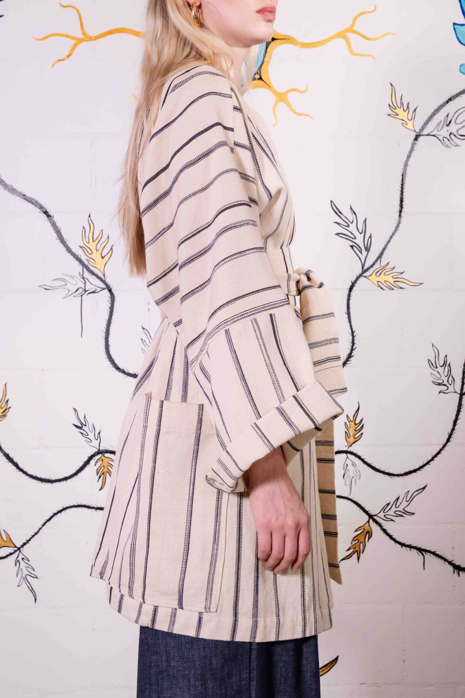 Model is wearing a midi-length, handwoven and hand-printed Benin cotton kimono by Atelier V.O, featuring traditional Dogon and Nagô-inspired patterns, a straight cut, drawstring closure, and wooden buttons.