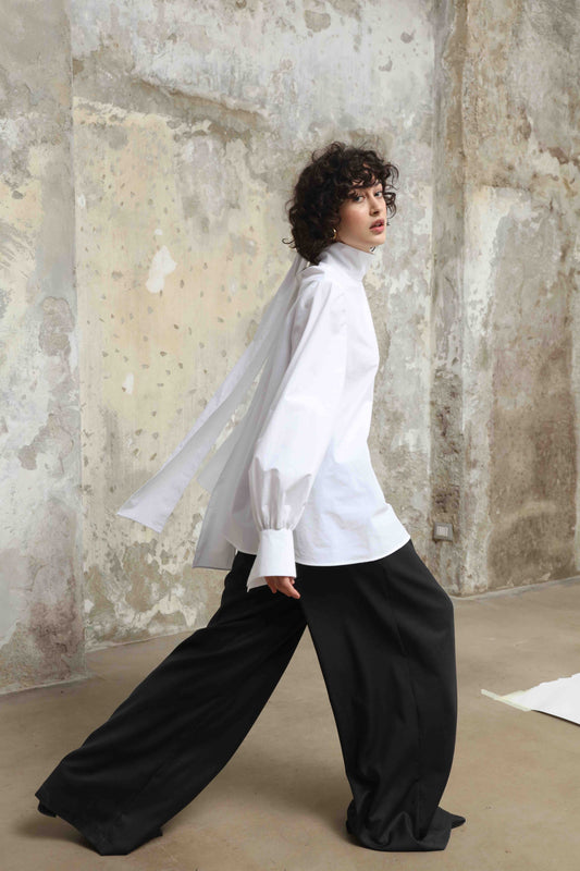 Regula Blouse by House of Woven Stories, handcrafted in Italy from 100% organic cotton poplin. Features adjustable long bow, minimalist tailoring, and sustainable design inspired by global craftsmanship.