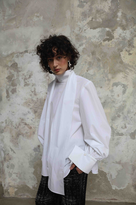 Regula Blouse by House of Woven Stories, handcrafted in Italy from 100% organic cotton poplin. Features adjustable long bow, minimalist tailoring, and sustainable design inspired by global craftsmanship.